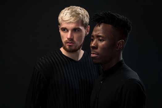 Portrait of two young african american and caucasian men standing over black background. International friendship concept. Studio shot