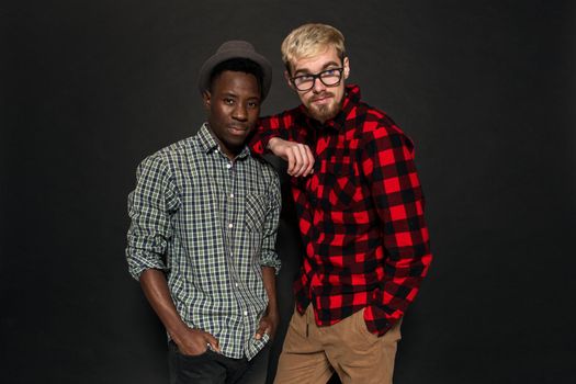 Studio shot of two stylish young men having fun. Handsome bearded hipster in a shirt in a cage standing next to his African-American friend in hat against a dark background. International friendship concept.