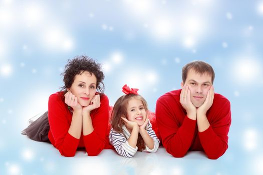 Happy young family dad mom and a little girl in bright red outfits . Family lying on the floor leaning on his hands.Blue Christmas festive background with white snowflakes.