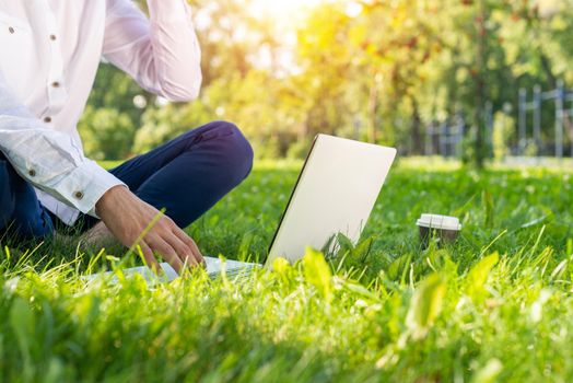 Businessman using laptop computer on green grass in park. Close up man hands typing on keyboard. Man in casual wear working with computer outdoors at sunny summer day. Freelance and blogging concept.