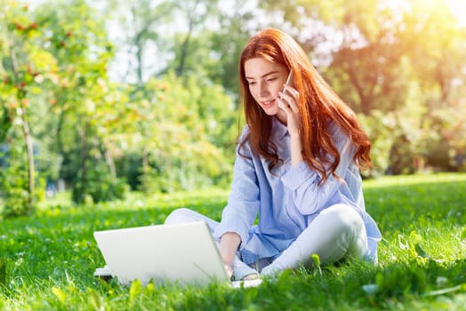 Young redhead woman sitting on green grass and using laptop. Charming girl talking on smartphone in park at sunny summer day. Mobile communication and conversation. Outdoors work and relaxation.