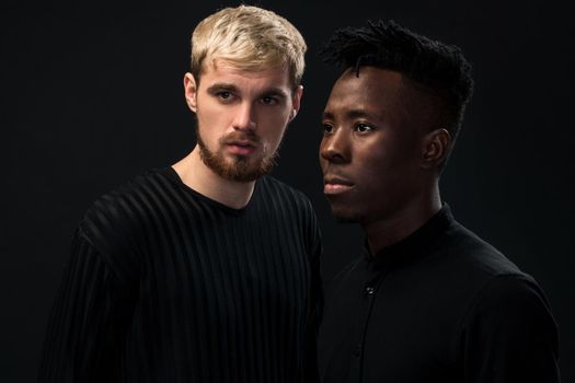 Portrait of two young african american and caucasian men standing over black background. International friendship concept. Studio shot