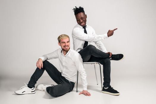 Joyful relaxed african and caucasian boys in white and black office clothes laughing and posing at white studio background with copy space. One man is sitting on the floor, the second is sitting on a chair