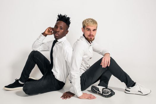 Friends. Two guys in white shirts and dark pants posing in the studio on a white background. Copy space. Two friends are sitting on the floor