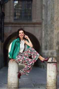 Young brunette woman, model of fashion, wearing green modern jacket and flower pants. Pretty caucasian girl with long wavy hairstyle smiling. Female with red lips in urban background.