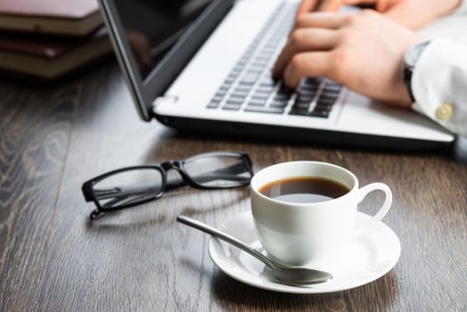 Businessman sitting at desk and working with laptop computer. Close-up of man hands typing on keyboard in office. Businessman workplace with glasses and cup of coffee. Innovations and entrepreneurship