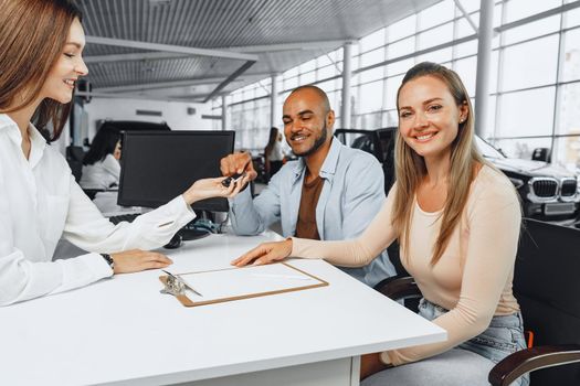 Cheerful couple taking keys of their new car in car dealership from saleswoman