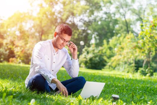 Young businessman sitting on green grass and using laptop computer. Handsome man working with computer in park at sunny summer day. Outdoors nature journey and relaxation. Freelance work concept.