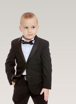 Beautiful little blond boy in a fashionable black suit with a tie.On a gray background.