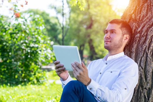 Dreamy man using tablet computer under tree in park on sunny day. Handsome businessman looking over tablet screen. Dreamy man sitting on green grass leaning against tree. Mobile communication concept