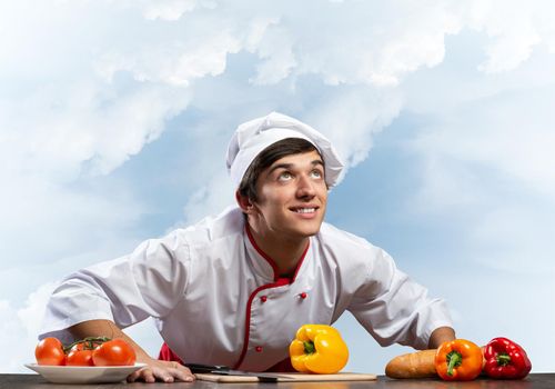 Young smiling chef standing near cooking table with fresh vegetables. Pleased chef in white hat and red apron on blue sky background. Cooking classes advertising. Restaurant food preparation concept