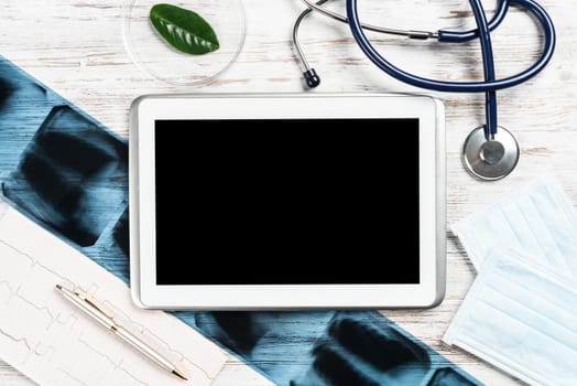Medical diagnostics in modern hospital. Top view tablet computer with blank screen, x-ray images, stethoscope and cardiogram on wooden desk. Professional healthcare and digital technology in clinic.