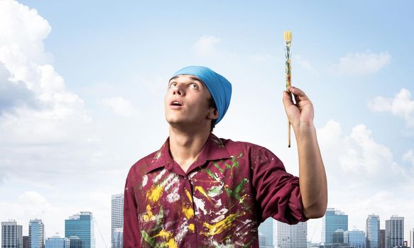 Young painter artist holding paint brush overhead. Portrait of decorator looking upwards on blue sky background. Artistic occupation and creative profession. Art school student posing.