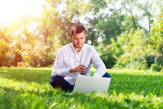 Young businessman sitting on green grass and using laptop computer. Handsome man working with computer in park at sunny summer day. Outdoors nature journey and relaxation. Freelance work concept.