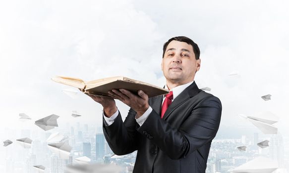 Serious businessman holding open book. Man in business suit standing on cloudy cityscape background with flying paper planes. Lawyer reading legal regulation book. Business accounting and consulting