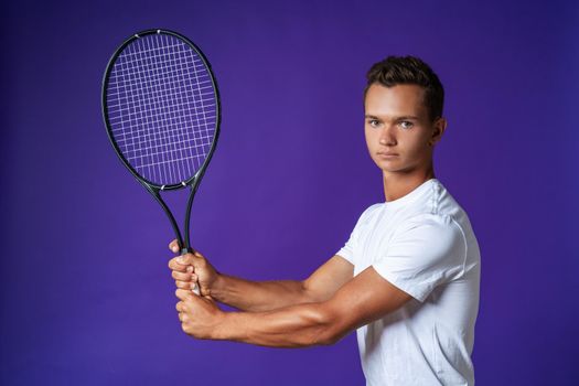 Caucasian young man tennis player posing with tennis racket against purple background close up