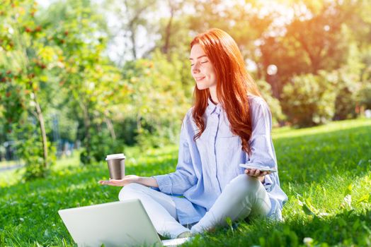 Young redhead woman meditating in lotus pose with laptop and cup of coffee on green grass. Beautiful girl in casual wear practicing of yoga with closed eyes. Outdoors work and relaxation at summer day