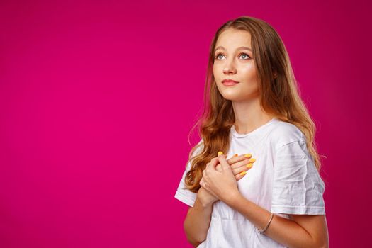 Young casual woman holding hands on her chest and looking up hopefully against pink background