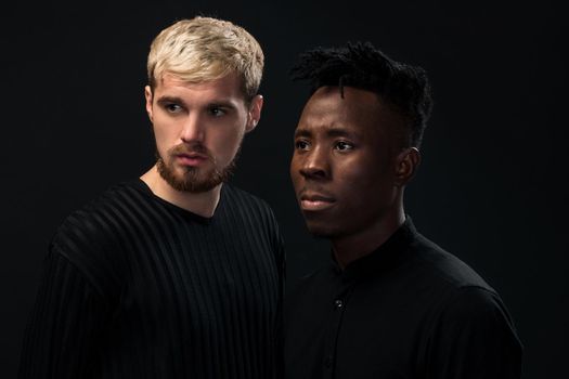 Portrait of two young african american and caucasian men standing over black background. International friendship concept. Studio shot