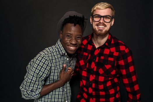 Studio shot of two stylish young men having fun. Handsome bearded hipster in a shirt in a cage standing next to his African-American friend in hat against a dark background. International friendship concept.