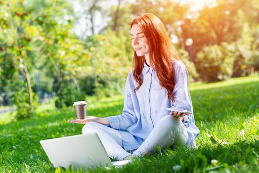 Young redhead woman meditating in lotus pose with laptop and cup of coffee on green grass. Beautiful girl in casual wear practicing of yoga with closed eyes. Outdoors work and relaxation at summer day