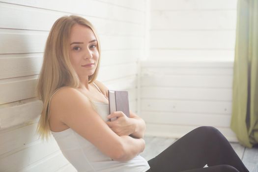 Lovely woman is sitting on the wooden floor with book over window.