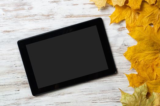 Tablet computer with blank screen lies on vintage wooden desk with bright foliage. Flat lay composition with autumn leaves on white wooden surface. Internet communication and digital technology