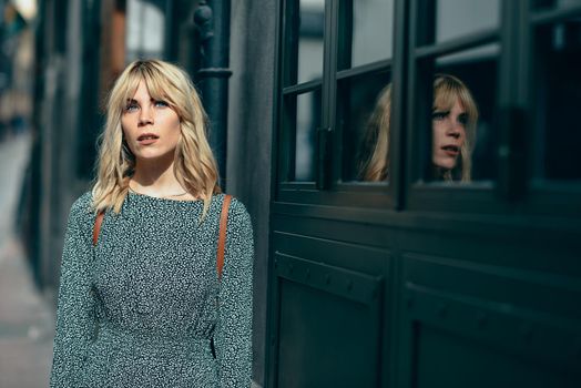 Portrait of serious young blonde woman standing on urban background. Beautiful girl with blue eyes wearing dress standing outdoors.