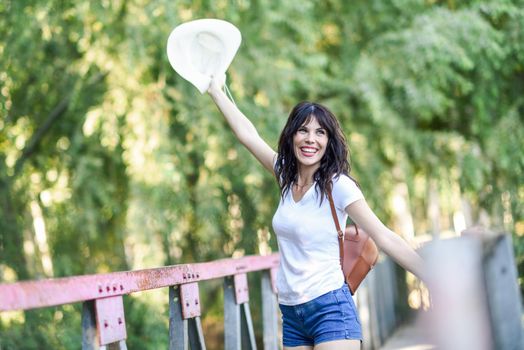 Beautiful hiker young woman with backpack, with blue eyes, wearing straw hat, hiking in the countryside and having fun on a rural bridge.