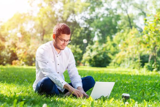 Young businessman sitting on green grass and using laptop computer. Handsome man working with computer in park at sunny summer day. Outdoors nature journey and relaxation. Freelance work concept.