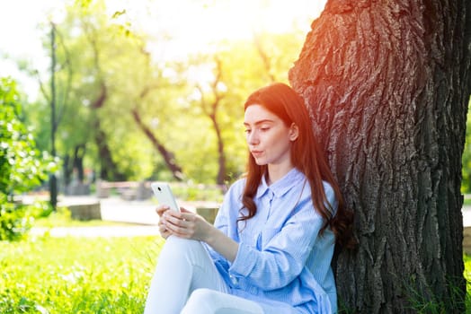 Young redhead woman using smartphone under tree in park on sunny day. Beautiful girl relaxing outdoors. Charming girl sitting on green grass leaning against tree. Healthy lifestyle and relaxation.