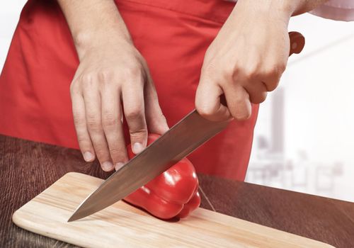 Chef hands cutting red fresh pepper on wooden board. Close up man hands cooking with knife on table in kitchen. Professional chef in red apron on light blurred background. Restaurant food preparation