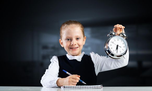 Smiling little girl holding alarm clock. Time to study and back to school concept. Happy schoolgirl sitting at desk with notebook and pen in hand. Time management. Research and education in school.