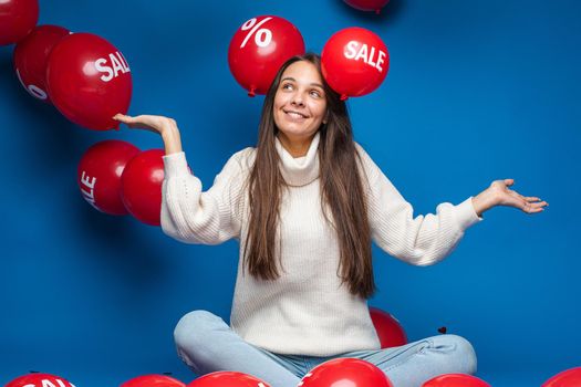 Pleased young woman sitting and looking on sale red air balloons, isolated on blue background. Shopping holiday concept