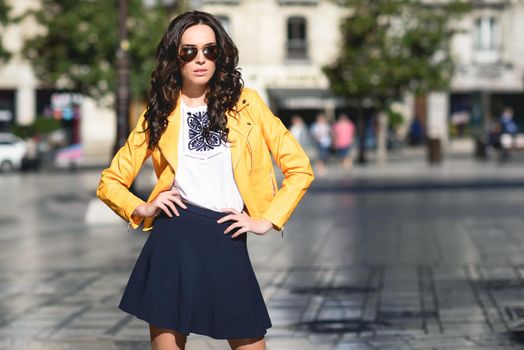 Young brunette woman with sunglasses. Girl, model of fashion, wearing orange modern jacket and blue skirt, standing in urban background.
