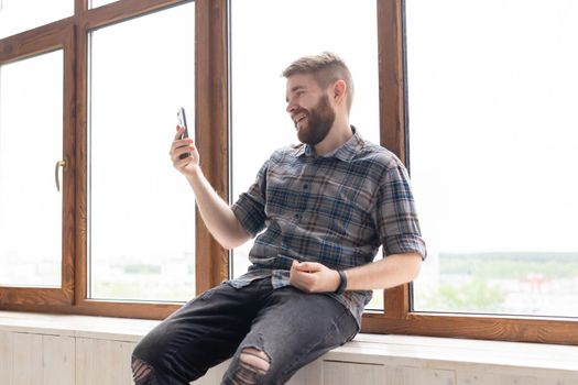 guy hipster with a beard is sitting on the windowsill talking on the video communication using a smartphone and wireless high-speed Internet.