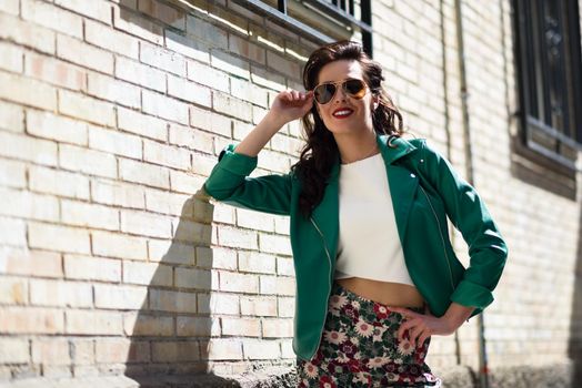 Young brunette woman with aviator sunglasses. Smiling girl, model of fashion, wearing green modern jacket, standing in urban background.