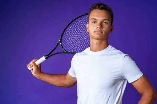 Caucasian young man tennis player posing with tennis racket against purple background close up