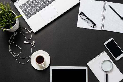 Top view of office workplace with digital gadgets. Flat lay black surface with laptop, green plant and cup of coffee. Top view coworking workspace with copy space. Digital technology and mobility.