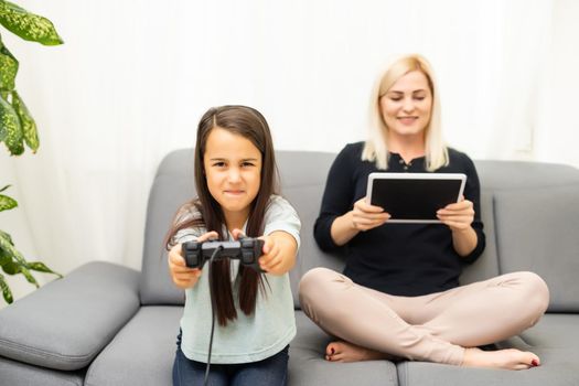 good relationship cute little girl with young mother using joystick playing video game sitting together in living room enjoying family holiday.