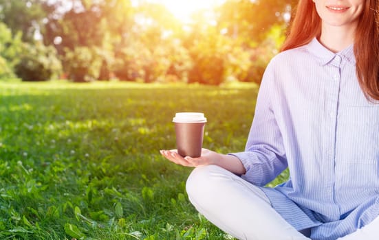 Young redhead woman meditating in lotus pose on green grass. Close up woman hand holding take away coffee cup. Girl in casual wear practicing of yoga in park. Mental relax and balance concept.