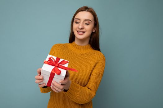 Photo shot of pretty positive surprised young dark blond woman isolated over colourful background wall wearing trendy outfit look holding gift box and looking at present box with red ribbon.