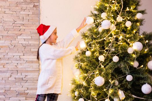 Little girl in sweater hanging ornaments on Christmas tree with light, bauble. Child decorating Xmas tree in beautiful family living room with fireplace.