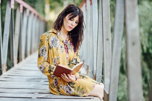 Young woman, wearing flowered dress, reading a book on a rural bridge.