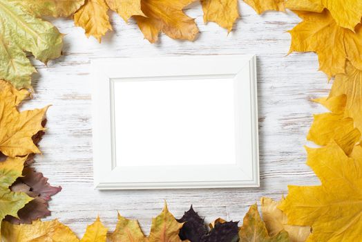 Blank rectangular photo frame lies on vintage wooden desk with bright autumn foliage. Flat lay with autumn leaves on white wooden surface. Simple white picture frame with copy space for design.