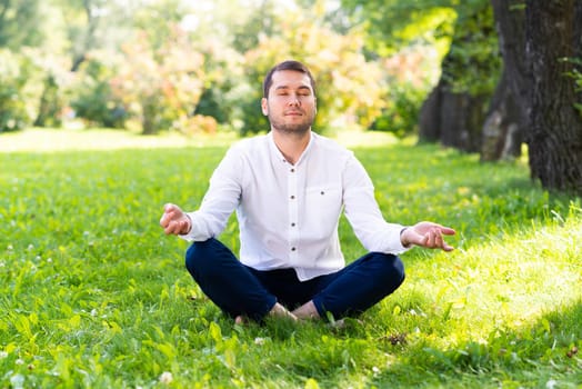 Young man meditates in lotus pose on green grass. Handsome man in casual wear practicing of yoga with closed eyes. Training and meditation outdoor at summer day. Healthy lifestyle and relaxation.