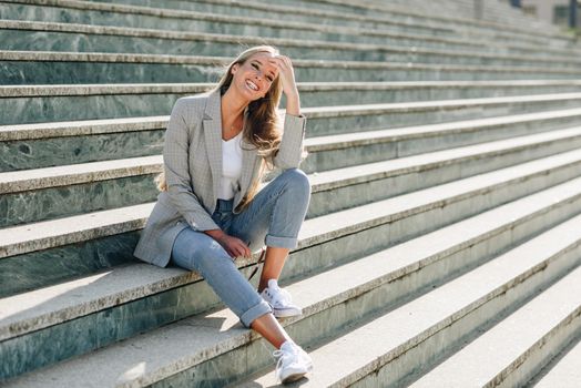 Beautiful young caucasian woman smiling in urban background. Blond girl wearing casual clothes in the street. Female with elegant jacket and blue jeans sitting on stairs.