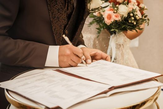 The groom's hand in a brown suit holds a pen and puts a signature on the consent of the marriage close-up.