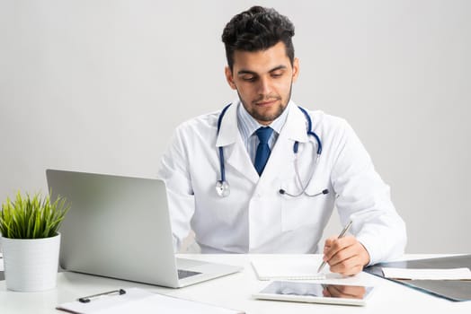 Handsome doctor sitting in office. Young practitioner in white coat with stethoscope at desk with laptop. Professional medical consultation and treatment in modern clinic. Medical healthcare concept