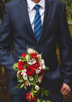 The groom in a white shirt, blue suit and tie holds the wedding bouquet of the bride of white and red flowers of roses.
